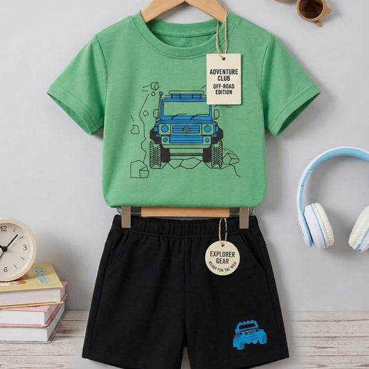 Children's green t-shirt with truck design and black shorts on a hanger, with books and sunglasses in the background.